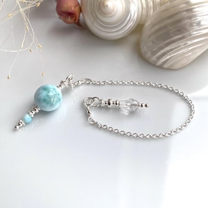 Puede incluir: Un péndulo plateado con una piedra veteada azul claro y blanca, conectado por una cadena a un cristal transparente. Este péndulo está diseñado para la adivinación y las prácticas espirituales.