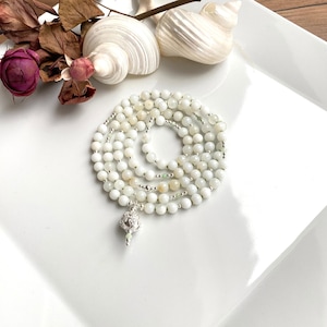 Può includere: Collana di perle bianche e crema con un ciondolo d'argento. La collana è avvolta su una superficie bianca.