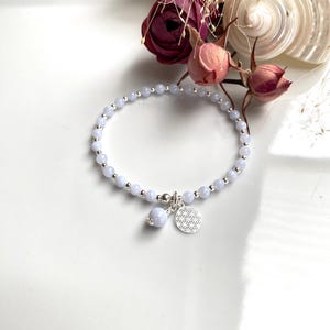 Könnte beinhalten: Ein zartes Armband aus blauen Achatperlen mit einem silbernen Anhänger mit geometrischem Muster. Das Armband liegt auf einer weißen Oberfläche.