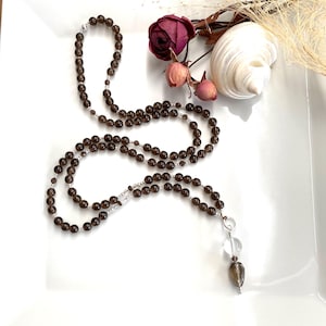 Op de afbeelding: Een lange, kralenketting met donkerbruine kralen en zilveren accenten. De ketting heeft een hanger van helder kristal en rookkwarts. De ketting wordt gepresenteerd op een wit oppervlak met gedroogde bloemen en een zeeschelp.
