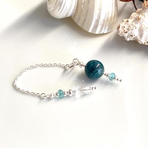 Puede incluir: Un collar de cadena de plata con un colgante de piedra preciosa azul verdoso y un dije de cristal. El collar está acentuado con piedras preciosas azul verdosas más pequeñas.