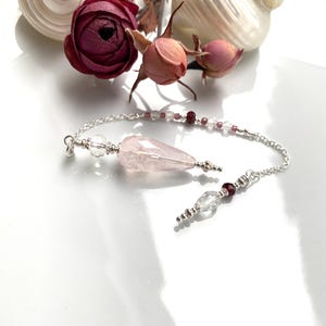 May include: A rose quartz pendulum with a silver chain, clear and red beads. The pendulum is teardrop-shaped. Dried roses and seashells are in the background.