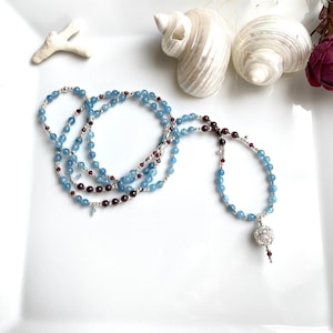 May include: A layered necklace with blue and red beads and a silver charm. The necklace is on a white surface with seashells.