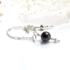 May include: A silver-toned pendulum with a black, spherical stone at the bottom. The pendulum features a chain, small clear beads, and a decorative top. The background is white with a few sprigs of dried grass.