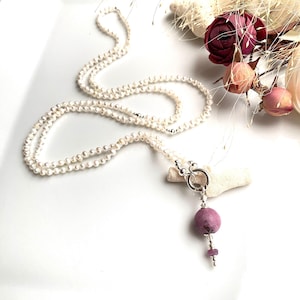 May include: A delicate pearl necklace with a silver clasp and a pendant featuring a pink gemstone. The necklace is draped on a white surface, with dried flowers in the background.