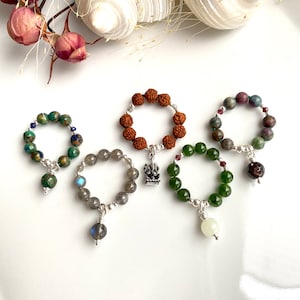 Puede incluir: Cinco anillos de cuentas con dijes. Los anillos están hechos de diferentes materiales, incluyendo cuentas verdes, marrones y grises. Los dijes son un elefante de plata, una esfera de plata, una esfera de plata, una esfera de plata y una esfera de plata.