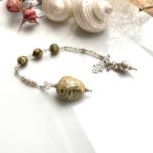 Könnte beinhalten: Ein silbernes Armband mit grünen und braunen Steinperlen und einem silbernen Anhänger. Der Anhänger ist eine Hand mit einem Loch in der Handfläche. Das Armband liegt auf einer weißen Oberfläche.