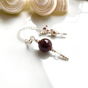 May include: A silver chain bracelet featuring a large, dark red gemstone bead and smaller clear and red beads. The bracelet is presented on a white surface, with two seashells visible in the background.