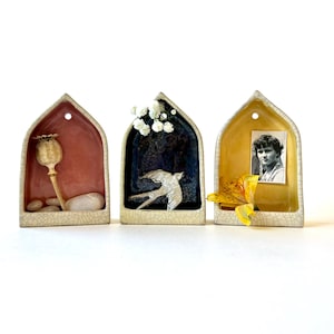 May include: Three ceramic wall shelves in the shape of arched windows, each with a different decorative element inside. The left shelf has a dried poppy seed pod and small stones, the middle shelf has a white bird, and the right shelf has a yellow flower and a black and white photo of a young woman.
