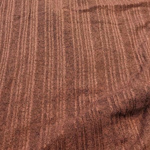 May include: Close-up of a textured fabric with vertical stripes in shades of brown and reddish-brown. The fabric has a slightly rough appearance, with visible variations in the weave. The overall effect is a warm, earthy tone.