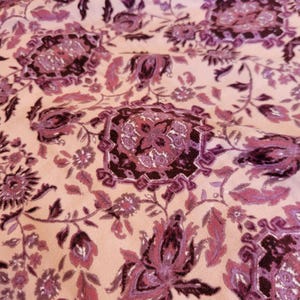 May include: Close-up of a fabric with a floral pattern. The design features large, stylized flowers and foliage in shades of purple and burgundy against a light pink background. The fabric has a soft, textured appearance.