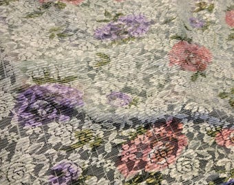 May include: Close-up of a floral lace fabric. The fabric features a repeating pattern of flowers in shades of pink, purple, and white, set against a light blue-green background. The lace has a textured, ribbed appearance.