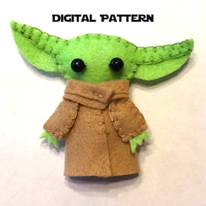Puede incluir: Un juguete de peluche verde de fieltro con grandes ojos negros y un abrigo de fieltro marrón. El juguete es un personaje de la franquicia Star Wars.