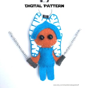 May include: A handmade felt keychain featuring a character with orange skin, blue clothing, and white and blue head tails. The character has black button eyes and is holding two silver pipe cleaner lightsabers. The words "DIGITAL PATTERN" are at the top.