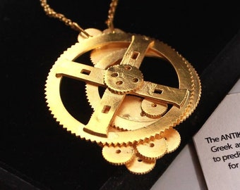Puede incluir: Un collar de oro con un colgante con forma de mecanismo de Anticitera, una antigua computadora analógica griega. El colgante está en una caja de terciopelo negro con una tarjeta blanca que dice "El mecanismo de Anticitera es una antigua computadora analógica griega y una omery utilizada para predecir las posiciones astronómicas y los eclipses para fines de calendario y astrológicos durante décadas por adelantado." La tarjeta también incluye el texto "Isla de Anticitera" y "Grecia".