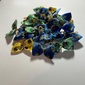 May include: A pile of small, colorful hair bows. The bows feature bee and floral patterns in shades of blue, yellow, and green. Each bow is tied with a yellow elastic band.