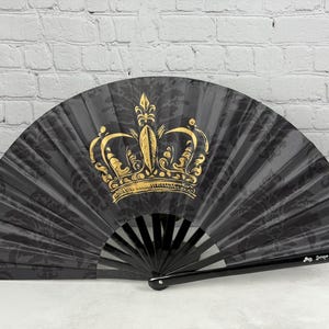May include: A black folding hand fan featuring a gold crown graphic in the centre and a subtle dark grey damask pattern across the fabric. The fan has black plastic ribs and the text Junque-Trunk.com printed on the lower right side.