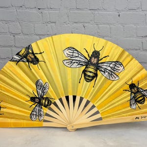 May include: A yellow folding hand fan featuring a repeating honeycomb pattern and four detailed illustrations of honeybees with black and yellow striped bodies and translucent wings. The fan has a light-coloured wooden frame and handle.