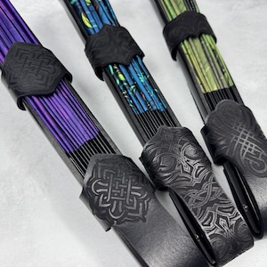 Op de afbeelding: Drie decoratieve handwaaiers met zwarte frames en ingewikkelde ontwerpen. Eén waaier heeft paarse stof, een andere blauwe en groene patronen en de derde groene en gele. Elke waaier heeft een zwarte leren riem met een Keltisch knoopontwerp.