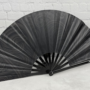 May include: A black hand fan featuring a subtle, dark grey swirling pattern across the fabric. The fan has a glossy black frame and a small white logo printed on the lower right side that reads Jacques-Prune.com in a clear, sans-serif font.
