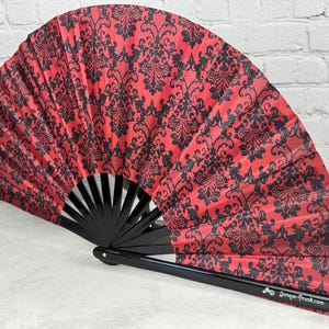 May include: A hand fan featuring a vibrant red fabric canopy with a repeating black damask pattern. The fan is constructed with black plastic ribs and a handle. The text Junque-Trunk.com is printed in white on the bottom right handle rib.