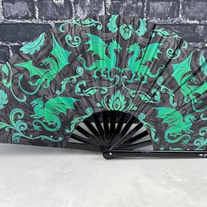 May include: A folding hand fan featuring a repeating pattern of bright teal dragons and swirling filigree motifs set against a dark charcoal grey background. The fan has black plastic ribs and a black handle with the text DragonFans.com printed on it.