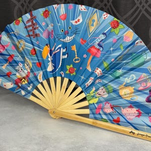 May include: A folding hand fan with a light blue background featuring a colorful Alice in Wonderland print. The design includes characters like the Cheshire Cat, the White Rabbit, and the Mad Hatter, alongside teapots, keys, and roses. The frame is bamboo.