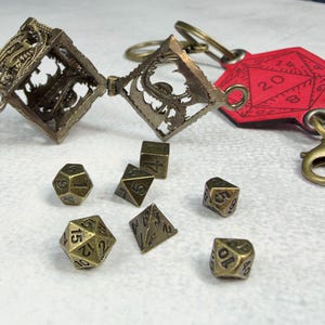 May include: A bronze-colored metal cage keychain with an openwork dragon design, attached to a red leather-like tag featuring a printed 20-sided die graphic. Six small bronze-colored polyhedral dice are arranged in front of the keychain.