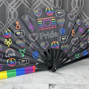 以下が含まれることがあります： 黒いPrideの扇子。虹色の縁があり、ハート、旗、平和のシンボルなど、さまざまなPrideのシンボルが描かれています。「Pride」の文字は虹色です。扇子は開いており、そのデザイン全体が表示されています。