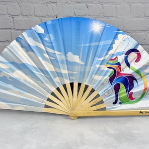 May include: A folding hand fan with a light wooden frame and a fabric leaf featuring a bright blue sky with white clouds and a sunburst. A stylised, multi-coloured cat illustration sits on the right side. The text Junque-trunk.com is printed.