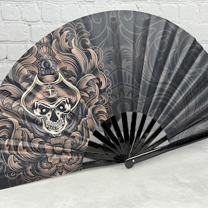 May include: A folding hand fan featuring a black and tan graphic design of a skull wearing a pirate hat with an anchor emblem, surrounded by ornate floral scrollwork. The fan has black plastic ribs and the text "Jumbo-Fan.com" printed on the handle.