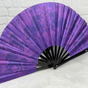May include: A purple hand fan with a black wooden frame. The fan features a repeating pattern of unicorns in shades of purple. The fan is open, displaying the full design. The text "Ginger-Peach.com" is visible on the fan.