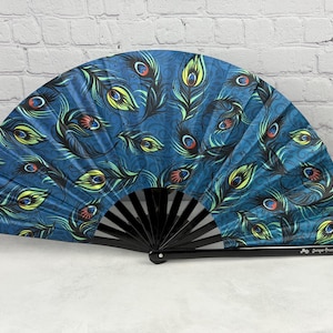 May include: A handheld folding fan featuring a deep blue fabric canopy decorated with a repeating pattern of peacock feathers in shades of green, yellow, and orange. The fan has black plastic ribs and a small logo printed on the handle.