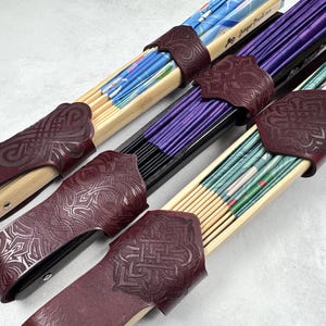 May include: Three folding hand fans with light wood or black frames, each secured by a dark burgundy leather sleeve featuring an embossed geometric knot pattern. The fans display blue, purple, and green patterned fabric leaves respectively.