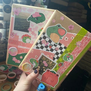 May include: Two brown journals decorated with pink and green frog stickers, pink flowers, and checkered patterns. The journals are covered in washi tape and have a variety of other decorative elements.