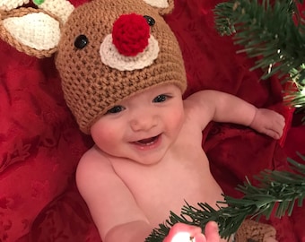 May include: A baby wearing a brown crocheted reindeer hat with a red pom-pom and a matching brown crocheted diaper cover. The baby is reaching for a lit Christmas tree light.