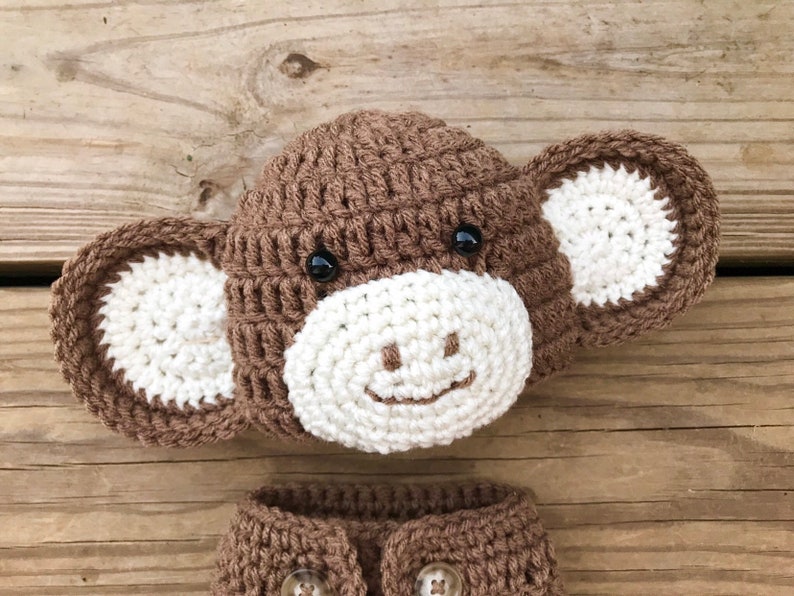 May include: A brown and white crocheted monkey hat and matching booties. The hat has a white face with black eyes and a smiling mouth.