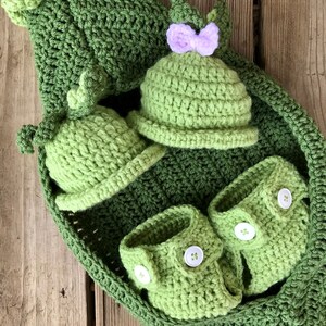 May include: A set of crocheted baby clothes in green, including a hat, booties, and a pea pod-shaped blanket. The hat has a purple bow.