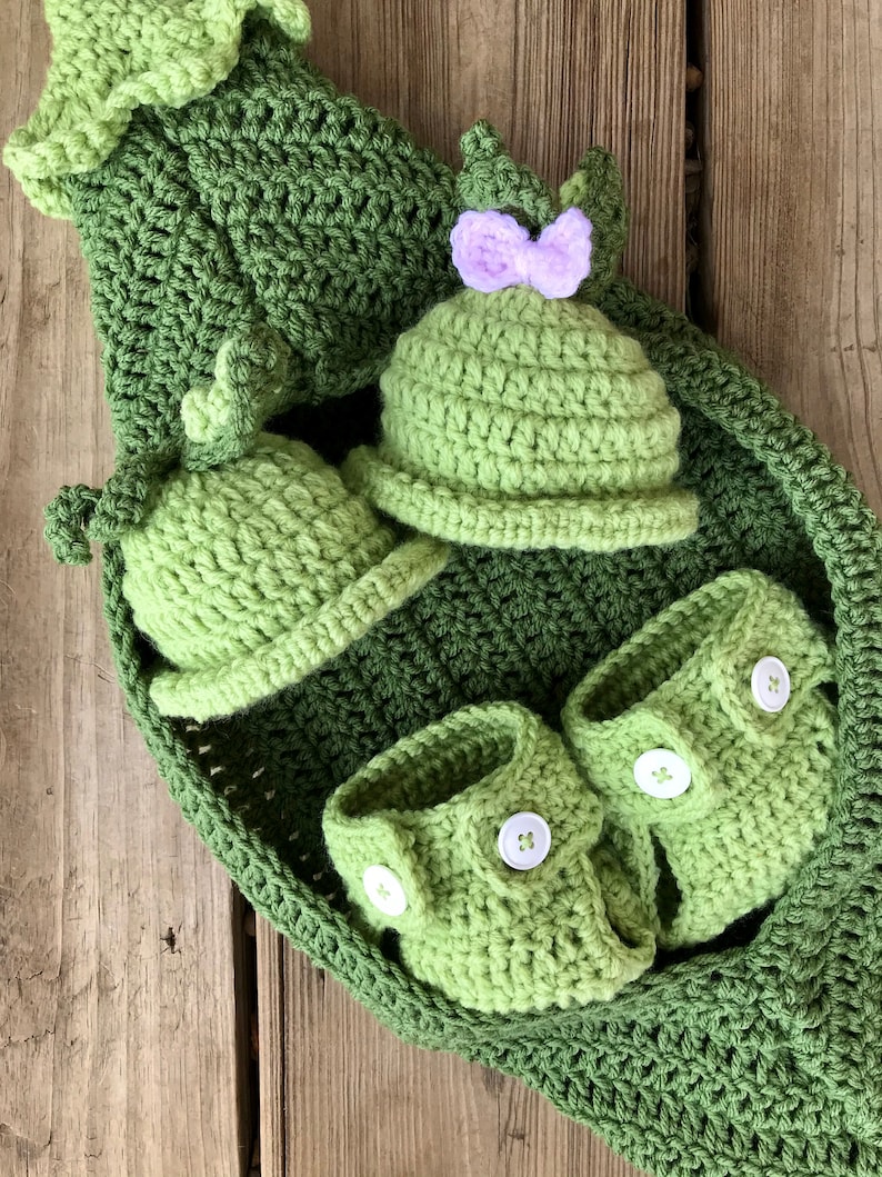May include: A set of crocheted baby clothes in green, including a hat, booties, and a pea pod-shaped blanket. The hat has a purple bow.