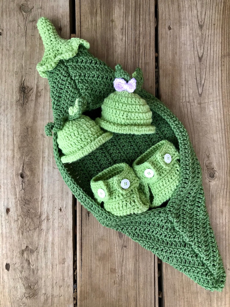 May include: A green crocheted pea pod with a green crocheted hat, two green crocheted booties, and a green crocheted pea pod hat with a purple bow.
