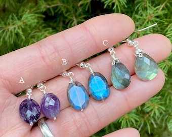 May include: A selection of gemstone earrings, each with a unique faceted teardrop shape. The earrings include amethyst, blue labradorite, and gray-green labradorite, all suspended from silver-colored ear wires. The background is a blurred view of green foliage.