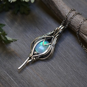 May include: A silver pendant necklace featuring a wire-wrapped design. The pendant holds a round, iridescent stone with shades of blue and green. The necklace is displayed on a wooden branch against a wooden background, with green foliage in the background.