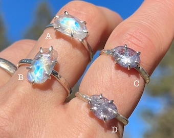 May include: Four silver rings with different gemstones. The rings are on a person's finger. The gemstones are a rainbow moonstone, a clear moonstone, a purple amethyst, and a clear amethyst.