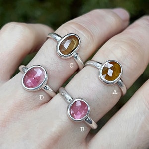 May include: Four silver rings with different colored gemstones. The rings are labeled A, B, C, and D. Ring A has a brown gemstone, ring B has a pink gemstone, ring C has a brown gemstone, and ring D has a pink gemstone.