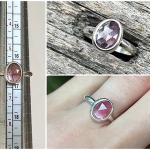 May include: A silver ring with a pink oval-shaped gemstone set in a bezel setting. The ring is shown on a finger and on a ring sizer.