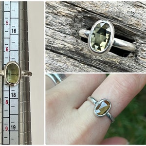 May include: A silver ring with a green oval gemstone. The ring is shown on a ring sizer and on a finger.