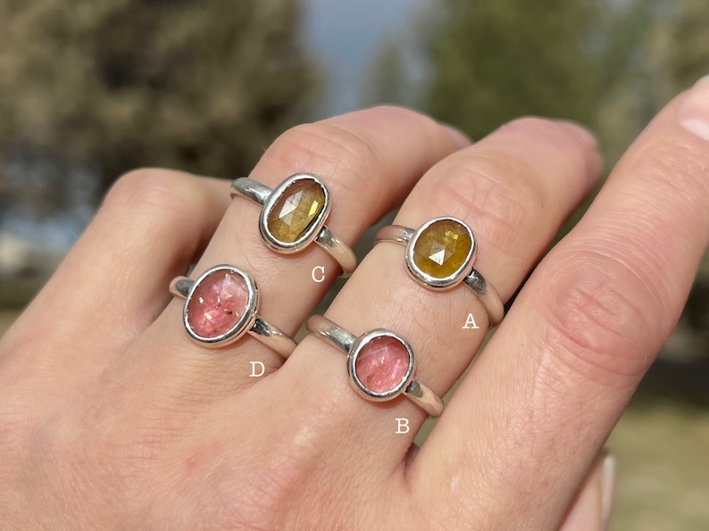 May include: Four silver rings with different colored gemstones. The rings are labeled A, B, C, and D. The gemstones are yellow, pink, and red.