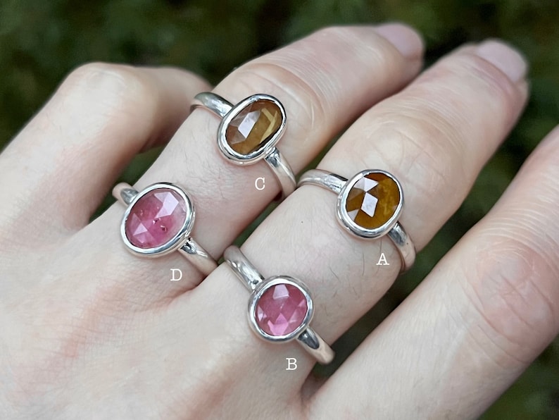May include: Four silver rings with different colored gemstones. The rings are labeled A, B, C, and D. Ring A has a brown gemstone, ring B has a pink gemstone, ring C has a brown gemstone, and ring D has a pink gemstone.