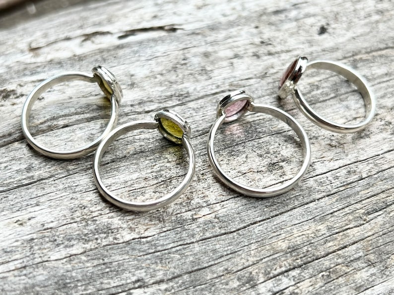 May include: Four silver rings with different colored gemstones. The rings are on a wooden surface.