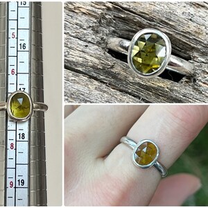 May include: A silver ring with a green oval gemstone. The ring is shown on a finger sizing tool and on a finger.